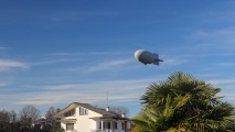 A blimp hovers above houses and a palm tree.