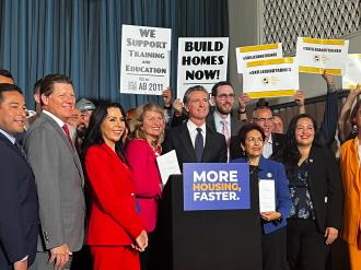California officials and construction workers gather at a podium with signs saying "More Homes Faster," "Build Homes Now," etc.
