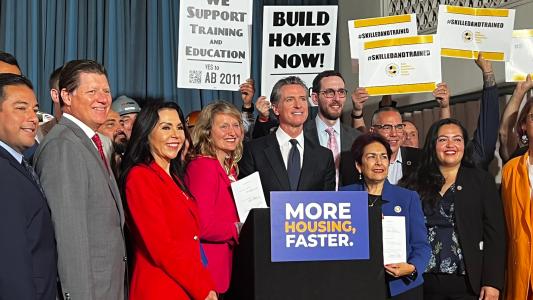 California officials and construction workers gather at a podium with signs saying "More Homes Faster," "Build Homes Now," etc.