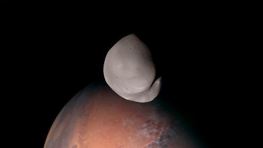 a grey, lumpy moon in front of the red surface of Mars