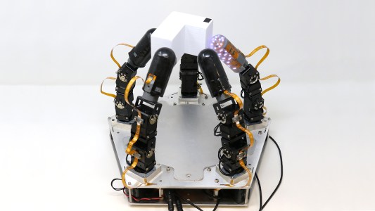 a robot hand comprising five fingers attached to a pentagon-shaped base. the fingers are holding an L-shaped block