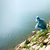 a woman in blue medical gear sitting on the edge of a lake.