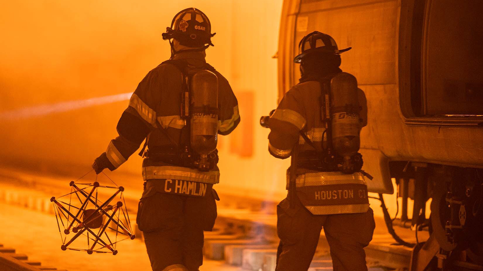two firefighters walking away from the camera. one is holding a robot made of rods and cables