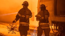 two firefighters walking away from the camera. one is holding a robot made of rods and cables