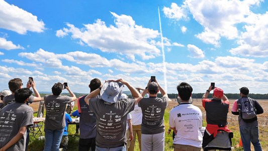a crowd of young people with their backs to the camera watching a rocket fly through the air in the distance