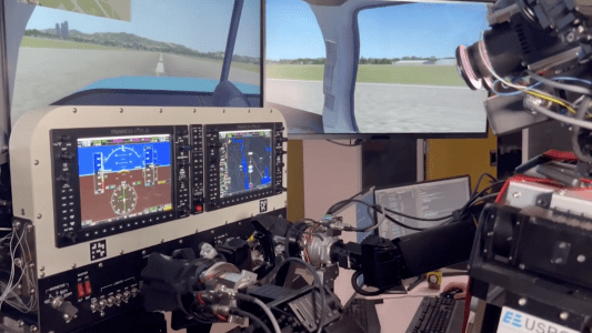 PIBOT, a humanoid robot pilot, is sitting in front of two monitors in a flight simulator.