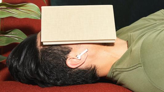A man lying down with a book opened over his face and earbuds in his ears