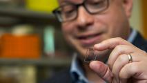 A man is holding up a living pharmacy prototype, which looks like a small piece of plastic with lines on it