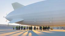 an airship outside a hangar with many people standing near it