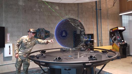 A man in camouflage is standing in front of a table with a virtual globe displayed above its surface