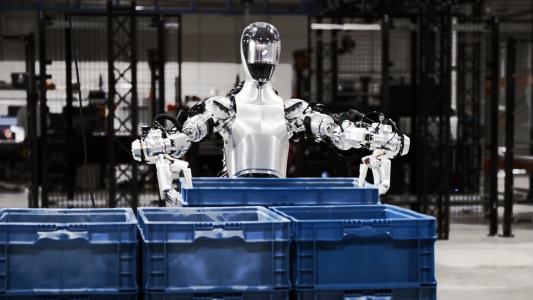 A humanoid robot grabs a blue crate off a stack in a warehouse