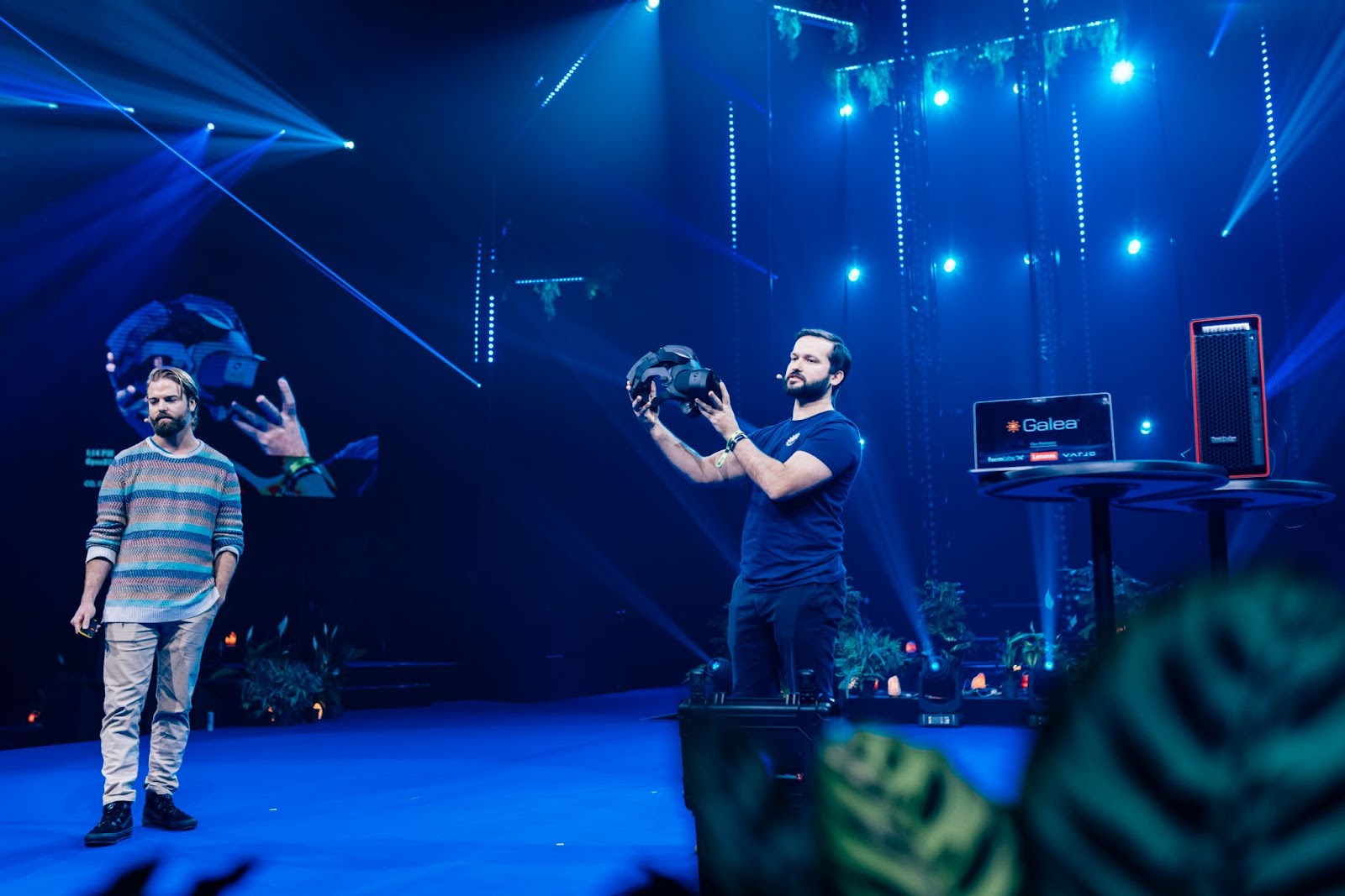 Two men standing on stage. One holds a black headset in his hands.