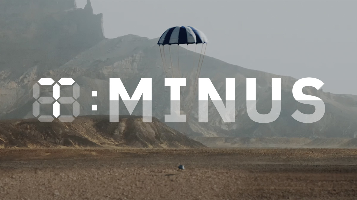 a capsule with a parachute attached to it landing in the desert. The T-Minus logo is on top of the image.