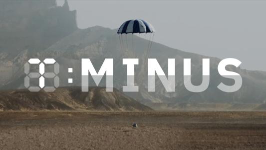 a capsule with a parachute attached to it landing in the desert. The T-Minus logo is on top of the image.