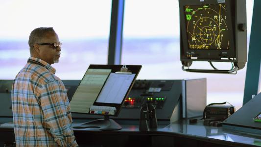 A man in a plaid shirt stands in front of an air traffic control monitor.