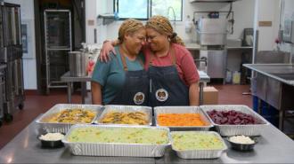Two women hugging in front of a kitchen filled with an abundance of food, portraying the significance of "you are what you eat.