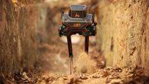 A bipedal robot walking through a ditch in a forested area, stirring up dust as it moves.