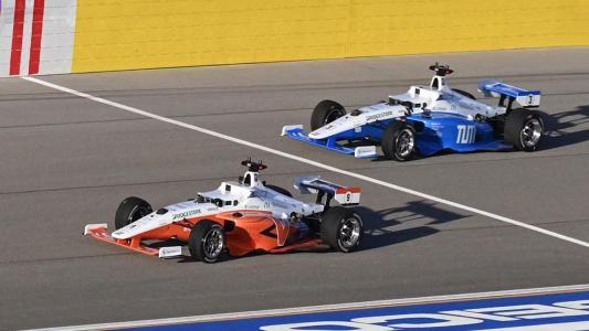 Two open-wheel race cars on a track, one orange and one blue, competing in an autonomous auto racing event.