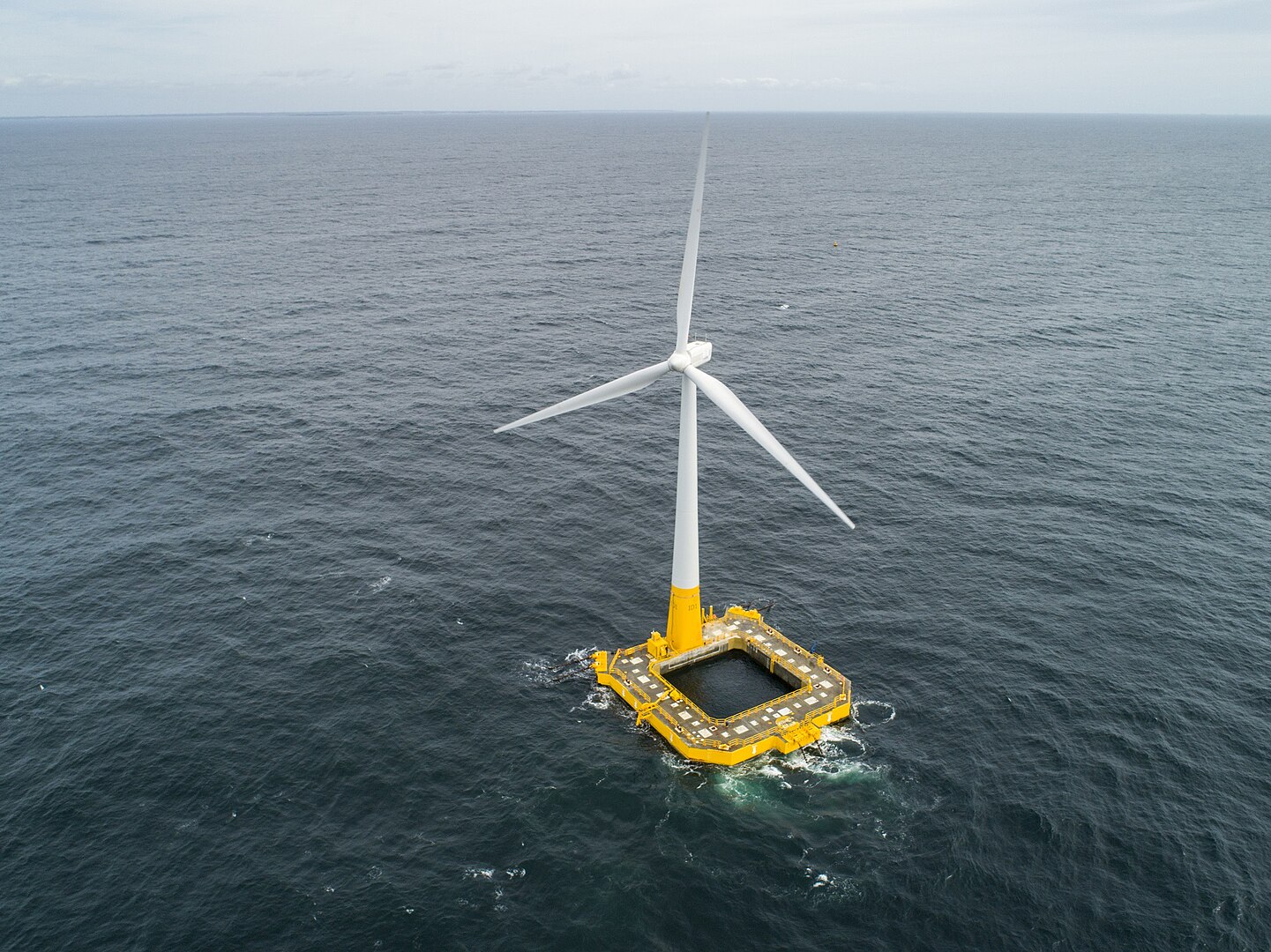 Aerial view of a solitary offshore wind turbine surrounded by open sea, with a cloudy sky above.