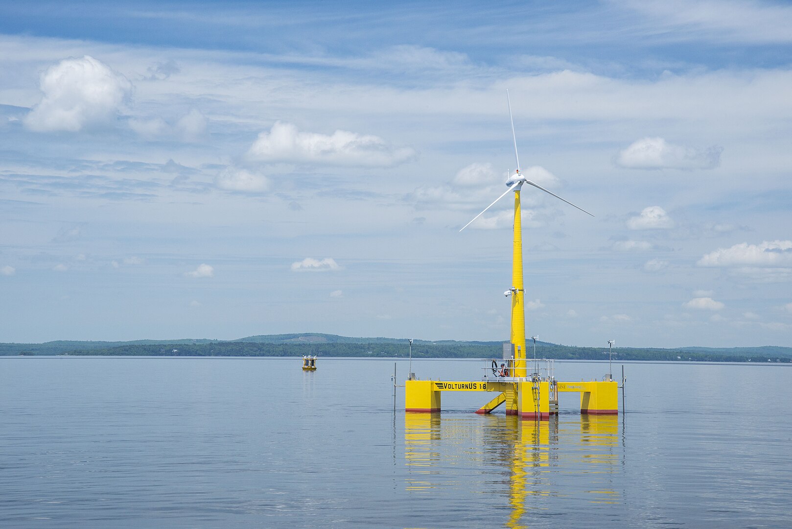A yellow floating wind turbine on a calm sea under a cloudy blue sky, with distant land visible on the horizon.