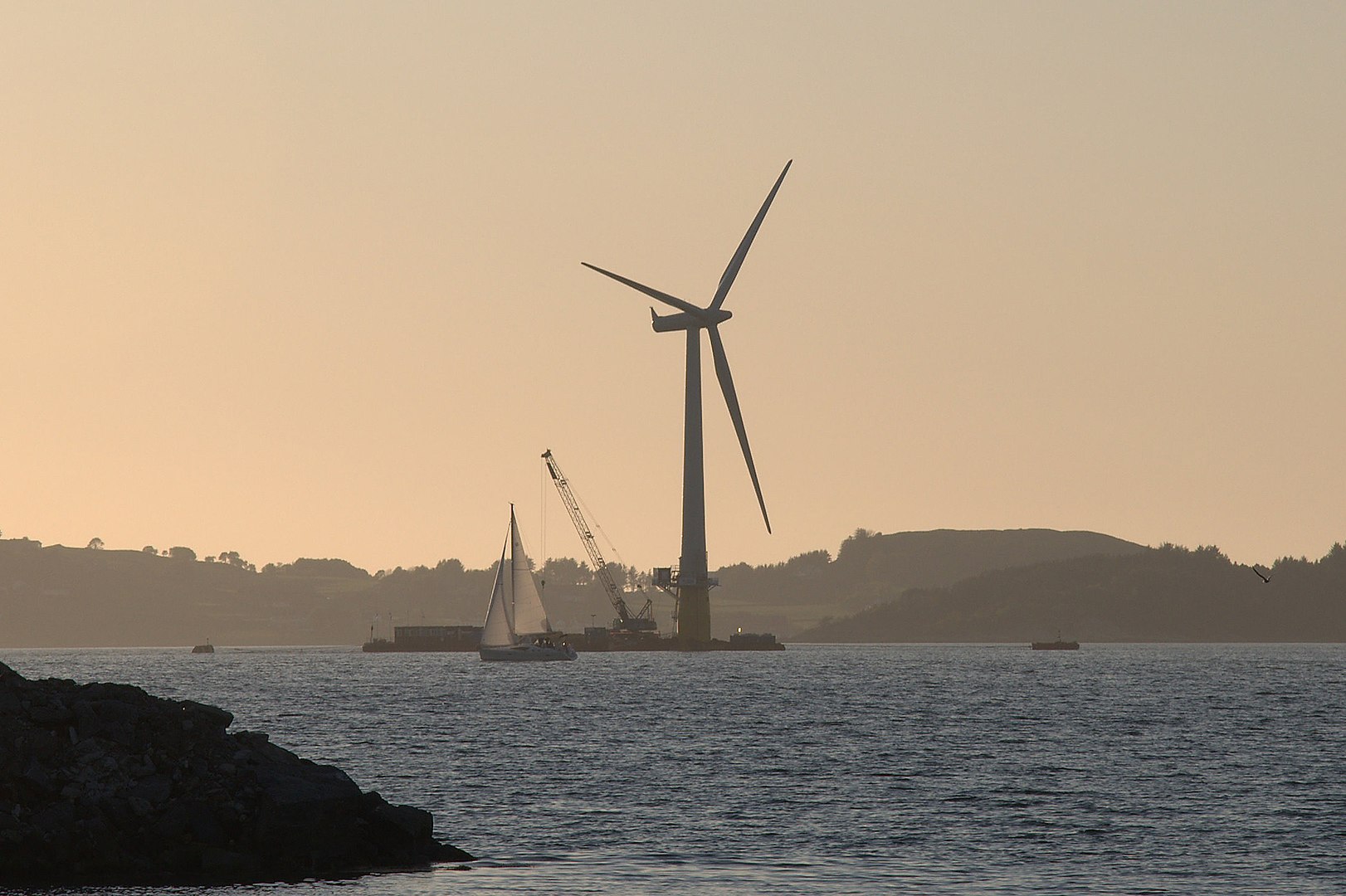 Silhouette of a wind turbine at dusk, with cranes and boats nearby, against a backdrop of a hilly coastline.