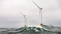 Two offshore wind turbines amid turbulent sea waves under a cloudy sky.