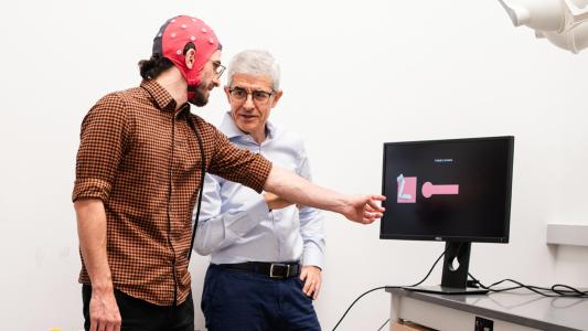 Two men standing near a computer monitor, with one man wearing an EEG cap and pointing at the screen and the other looking at him