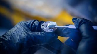 Gloved hands hold a small metallic component with tweezers under a blue light in a laboratory setting.