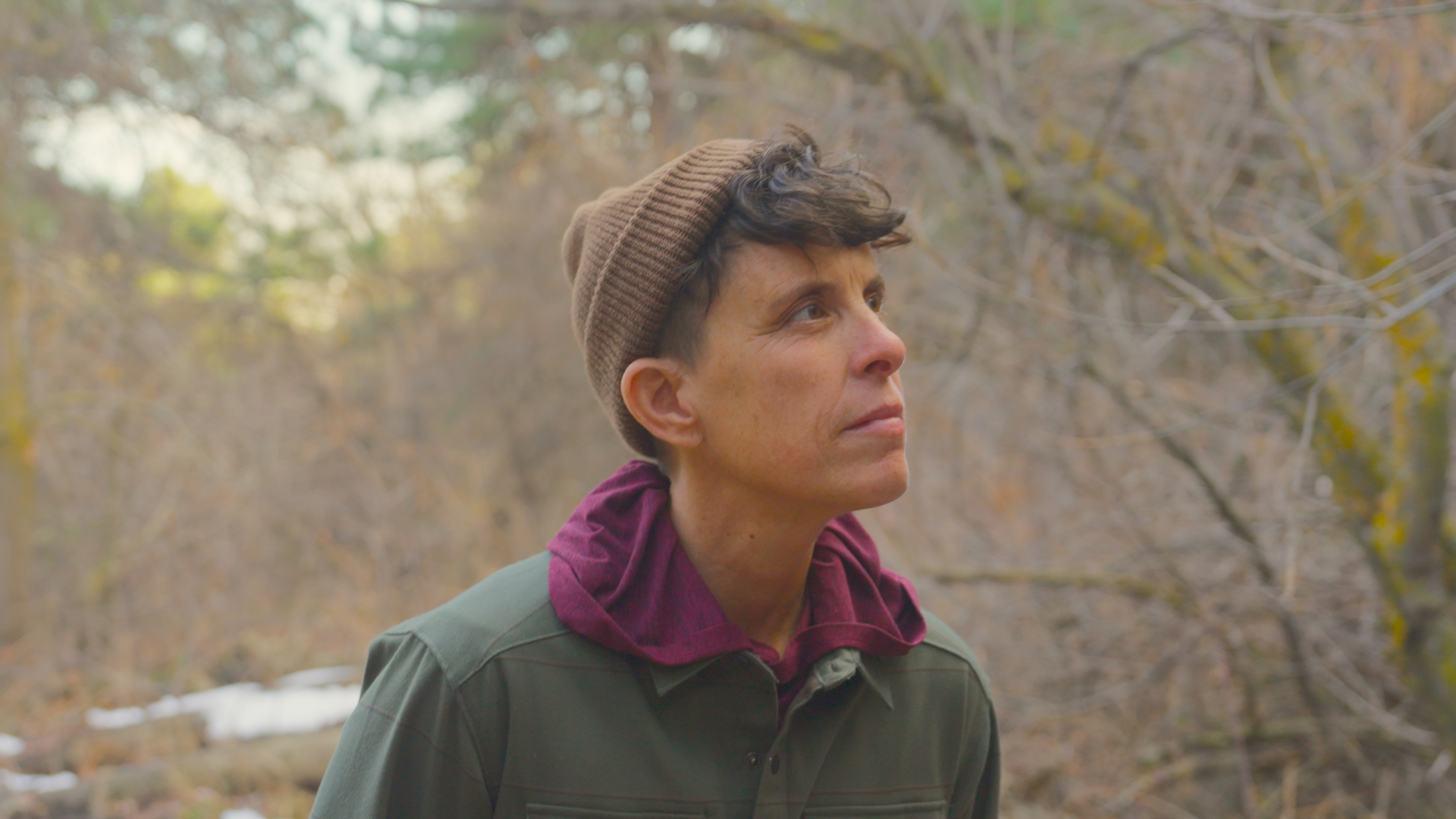 A person wearing a brown beanie and green jacket looks thoughtfully to the side outdoors in a wooded area.
