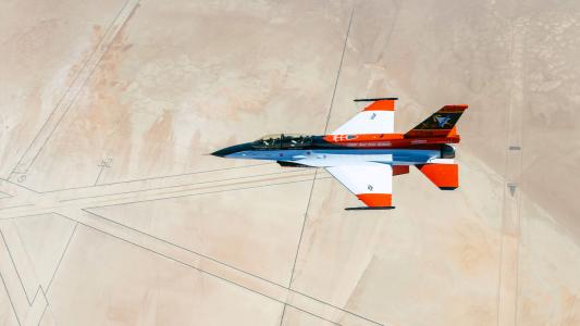 A view of an orange and blue jet in flight, with desert terrain visible in the background.
