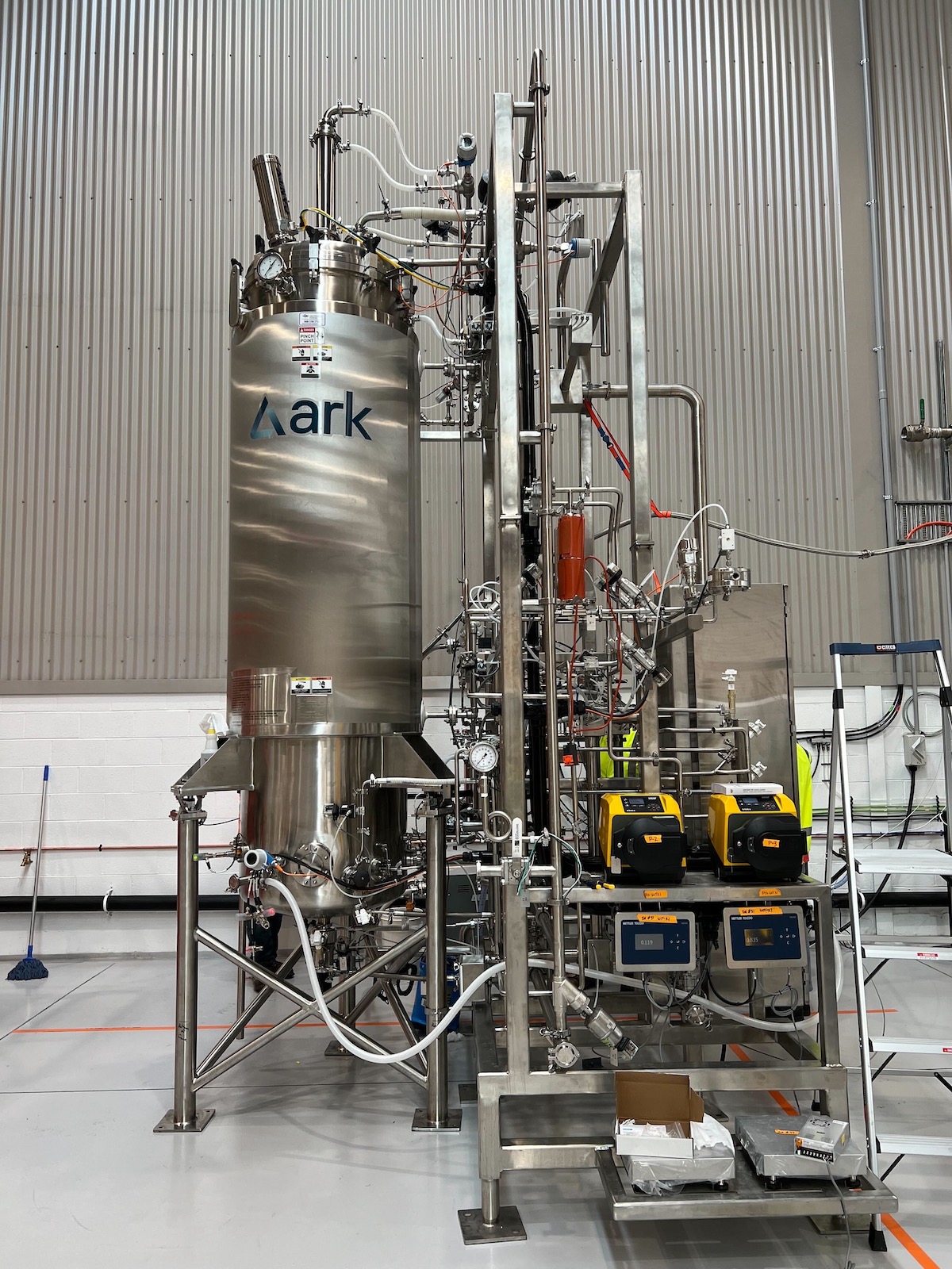 A large stainless steel bioreactor tank for cultivating meat, with various connected pipes and valves in an industrial facility.