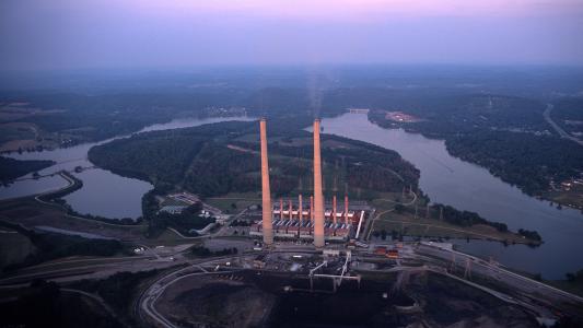 Aerial view of a large power plant with tall smokestacks near a river at dusk.