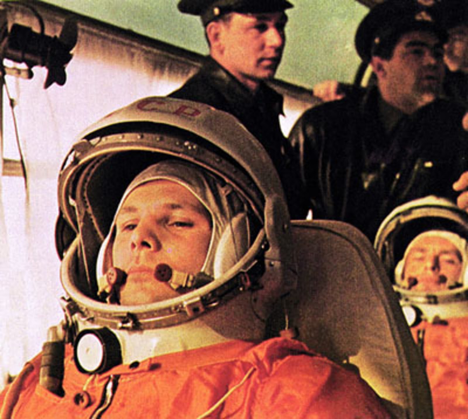 Two astronauts in full suits and helmets seated for a mission, with personnel in the background.