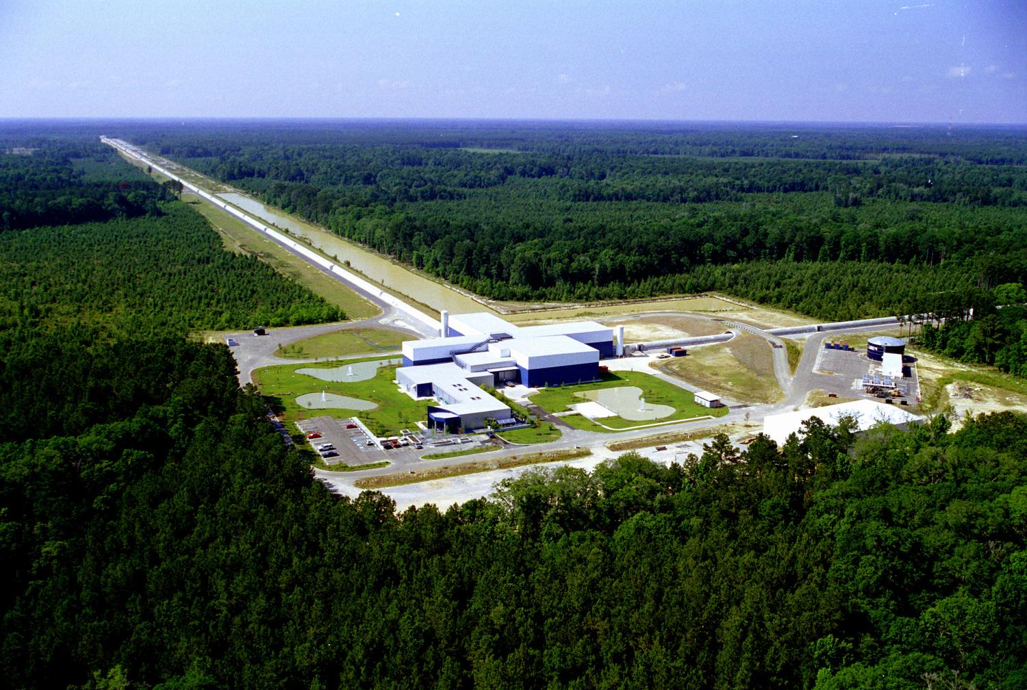 An aerial view of a building with two long tubes extending away from it perpendicular to one another. The entire facility is surrounded by woods.