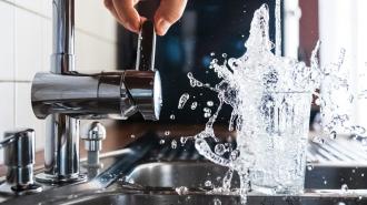 A person turning on a kitchen faucet, causing water to splash dramatically into a clear glass below.