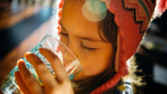 Young girl in a colorful knit hat drinking water from a clear glass, possibly containing forever chemicals, with sunlight creating a lens flare across the image.