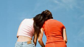 Two people standing close together, united by a surge of oxytocin, one wearing a white top and the other in a red top, against a clear blue sky.