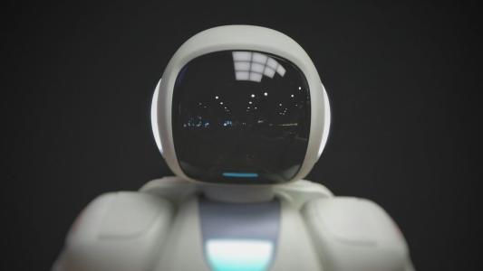 Close-up of a white humanoid robot with a reflective face screen and a blue light on its chest, set against a dark background.