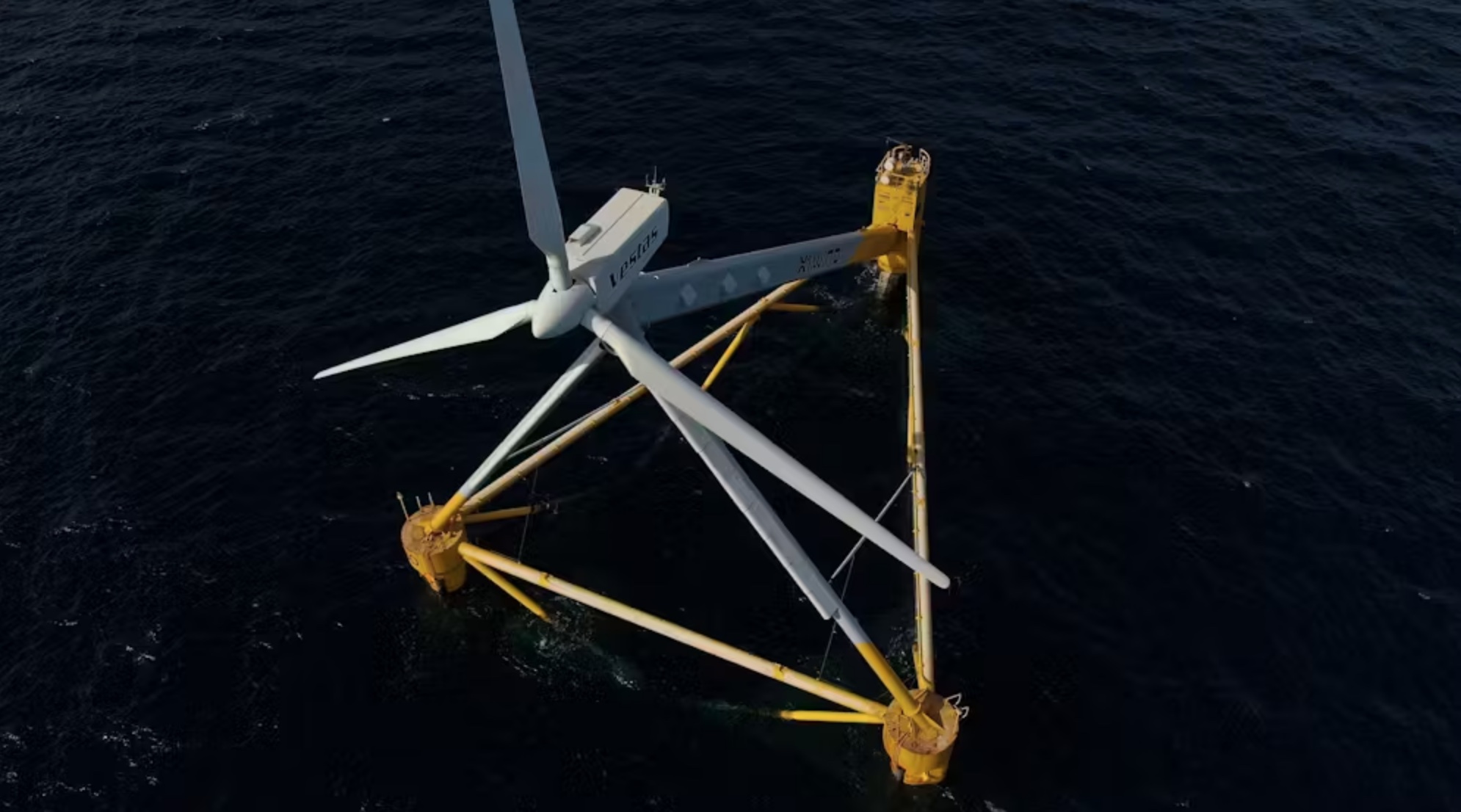 Aerial view of an offshore wind turbine with a white and yellow base standing in deep blue sea waters.
