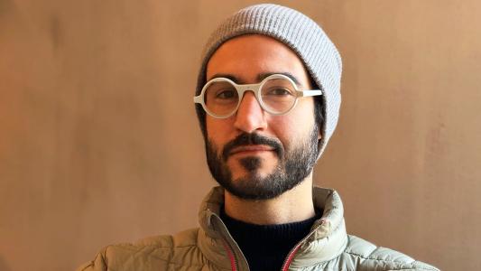 A man wearing a gray beanie, white AI glasses, and a green puffer jacket smiles subtly against a light brown background.