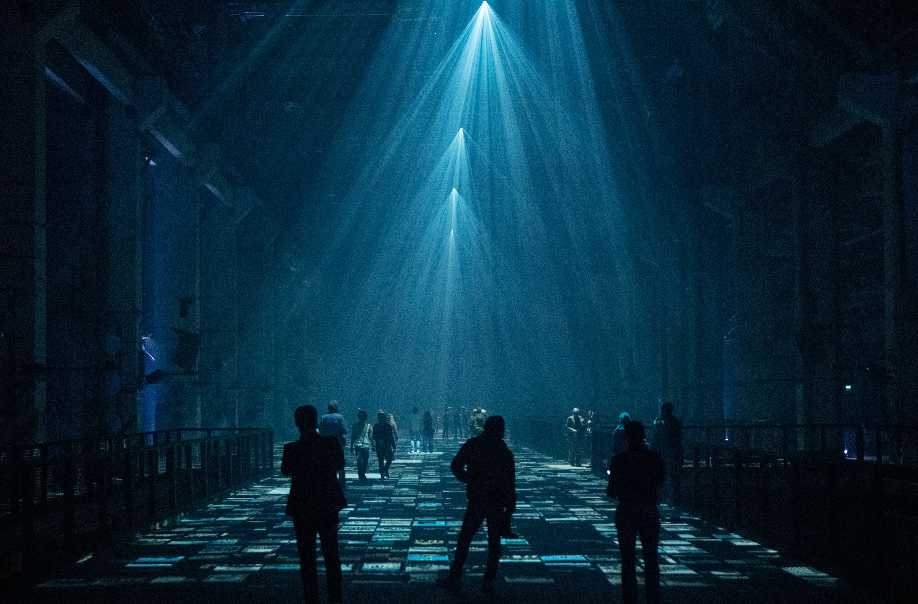 A group of people stands in a large, dimly lit industrial space with blue light beams shining down, creating geometric patterns on the floor.