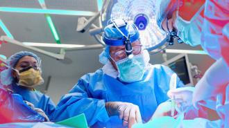 A surgeon in scrubs and a headlamp performs a gene-edited pig kidney transplant, assisted by another healthcare professional, under bright surgical lights.