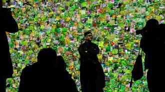 Man standing in front of a vibrant, colorful mosaic wall, being photographed by silhouetted figures in a dimly lit room.