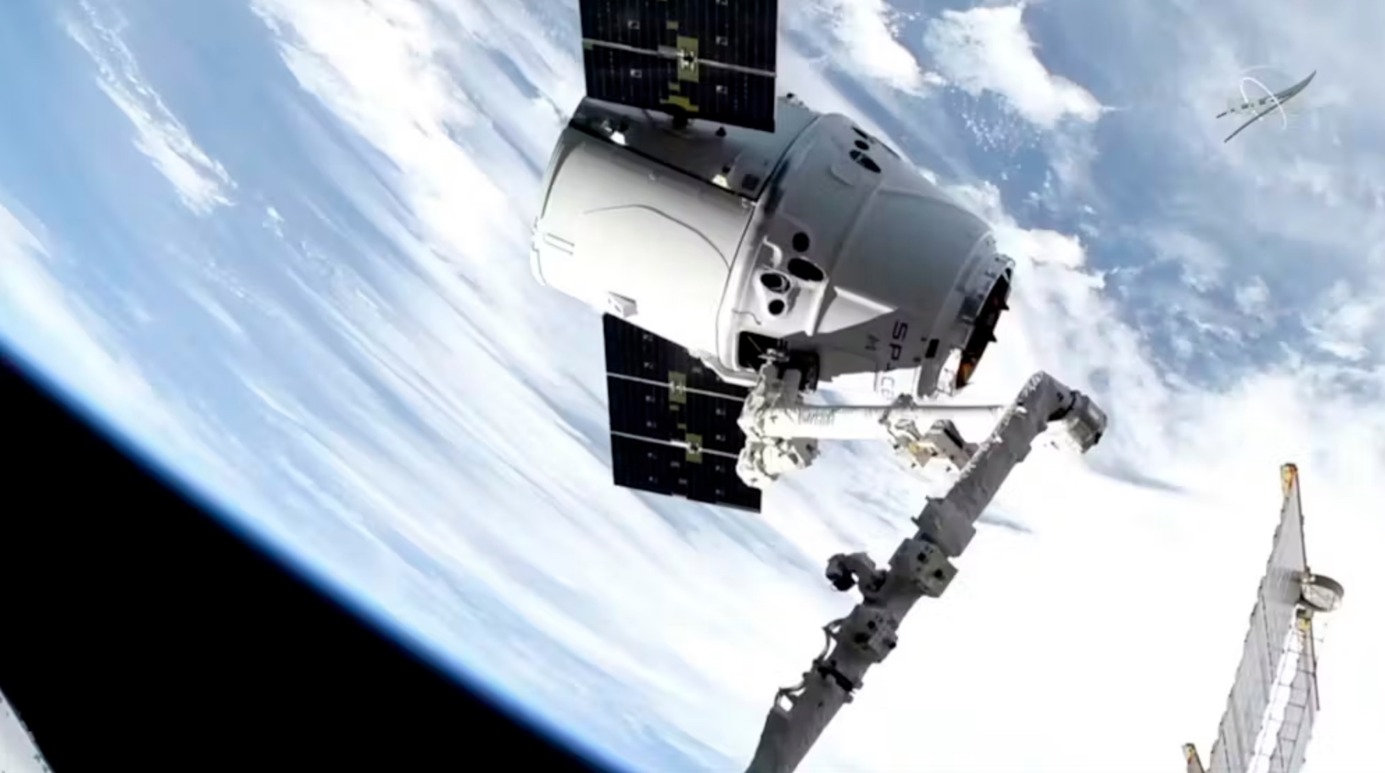 A spacecraft attached to a robotic arm above earth, with clouds and the curvature of the planet visible in the background.