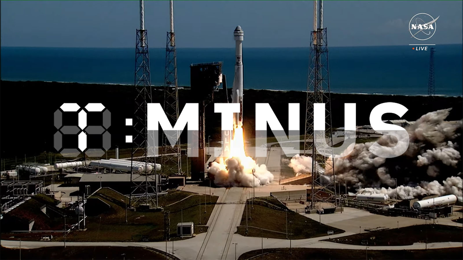 A rocket launches from a coastal launch pad, with "T-Minus" displayed prominently in front. The sky is clear with visible ocean in the background.