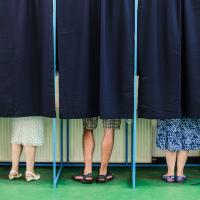 Six individuals standing behind closed blue curtains in voting booths, with only their legs and feet visible, suggesting they are engaged in the voting process.