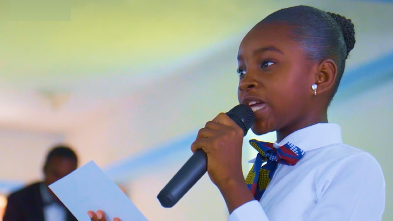 A young person in a school uniform speaks into a microphone while holding a piece of paper.