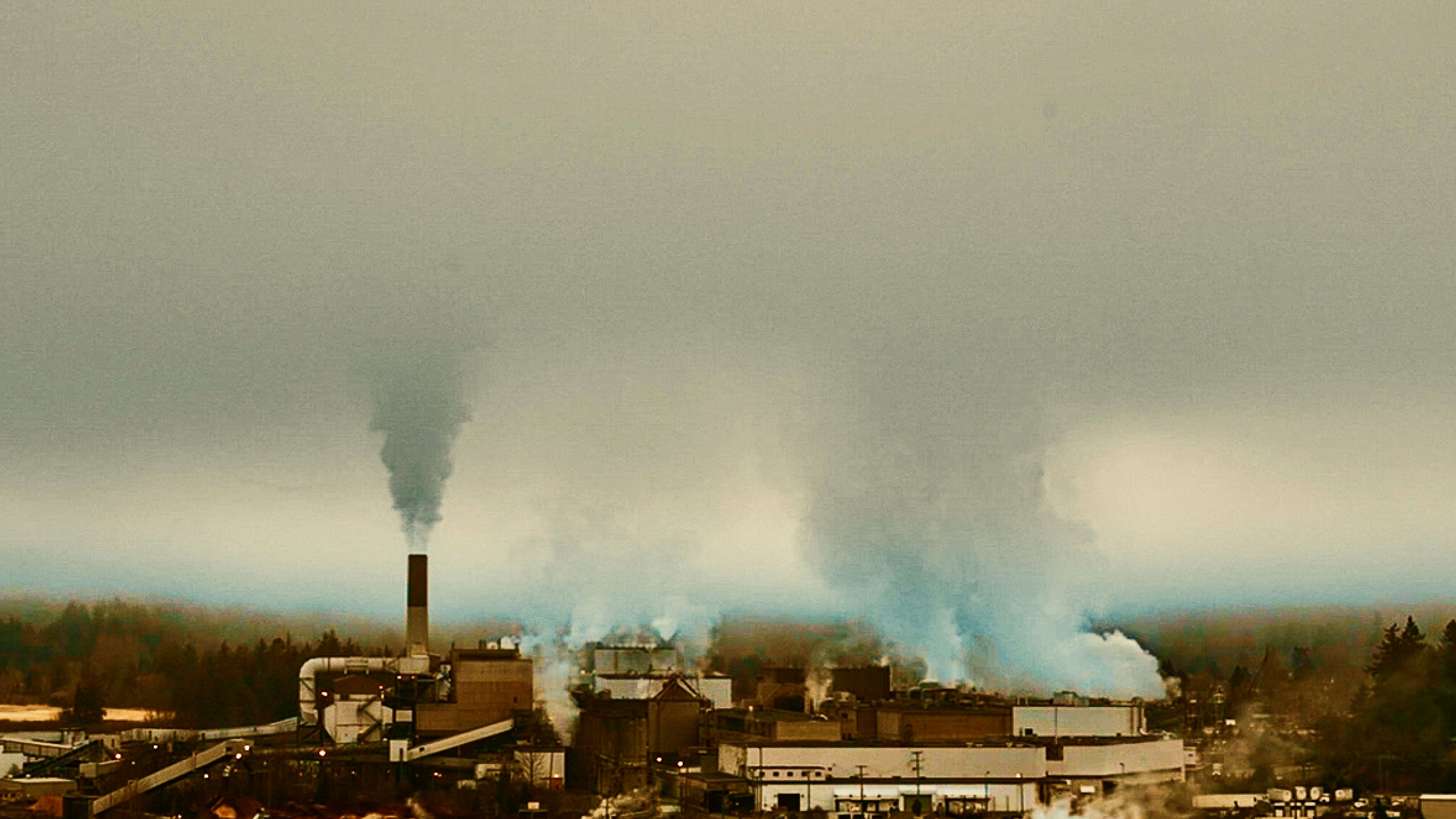 Industrial facility emitting smoke from multiple smokestacks under an overcast sky.