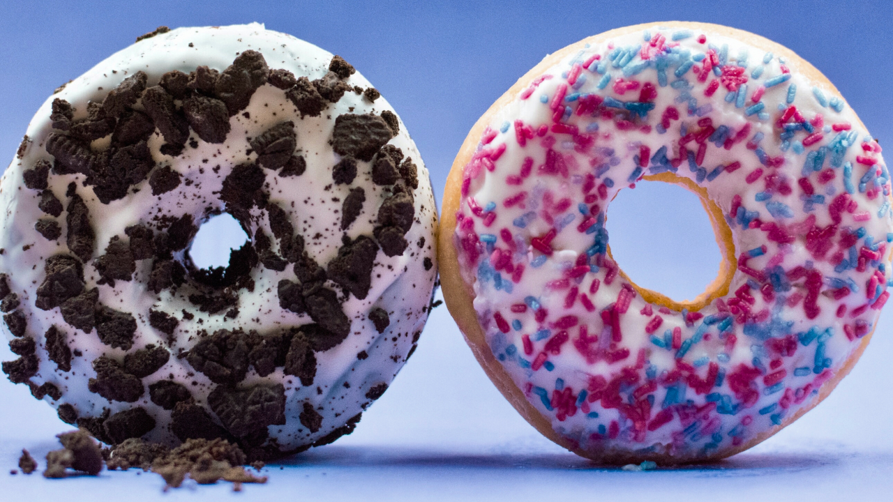 Two donuts with white icing on a blue background. The donut on the left is topped with cookie crumbles, and the one on the right is decorated with pink and blue sprinkles.