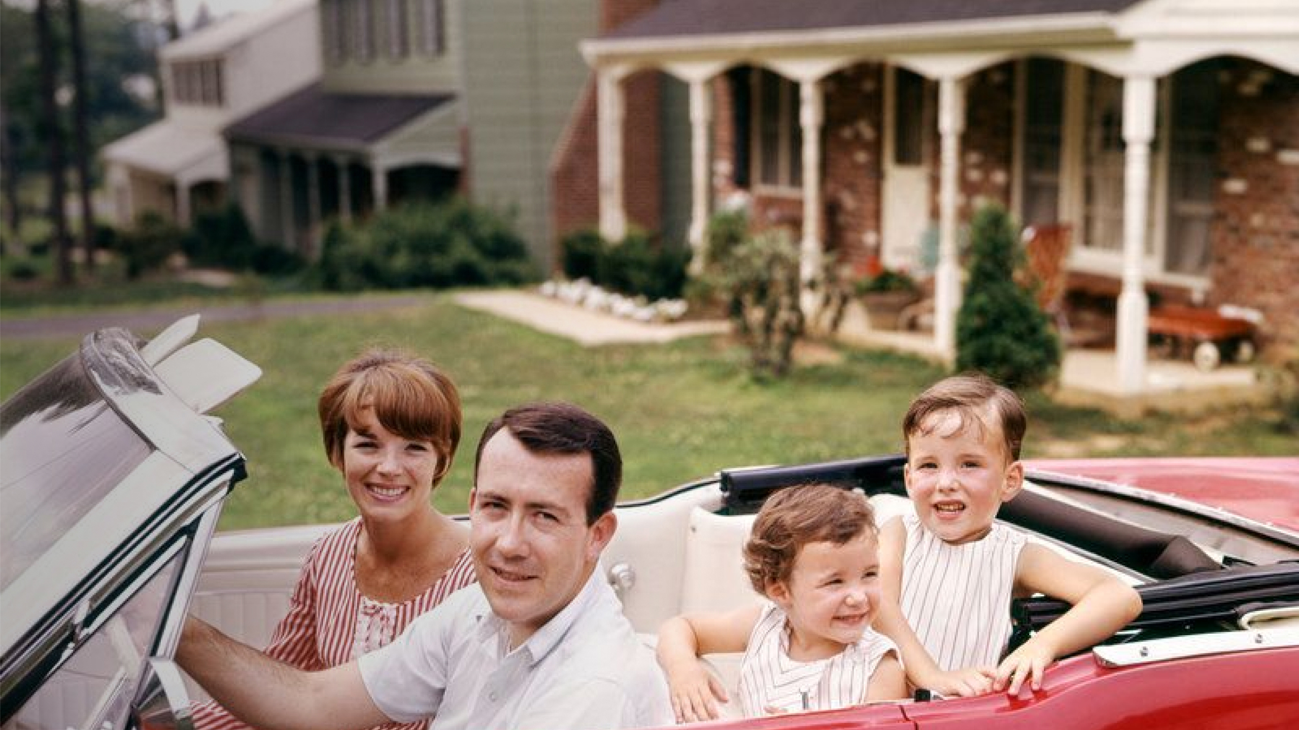 A family of four is seated in a red convertible car, parked in front of suburban houses. The parents sit in the front, while the two children sit in the back.